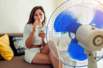Young woman cooling down by ventilator at home while drinking water and hanging in phone. Summer...