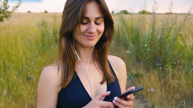 Happy young woman use smartphone and headphone standing in field at sunset, healthy lifestyle. Sportywoman runner using smartphone outdoor.