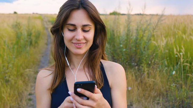 Happy young woman use smartphone and headphone standing in field at sunset, healthy lifestyle. Sportywoman runner using smartphone outdoor.