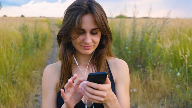 Happy young woman use smartphone and headphone standing in field at sunset, healthy lifestyle. Sportywoman runner using smartphone outdoor.