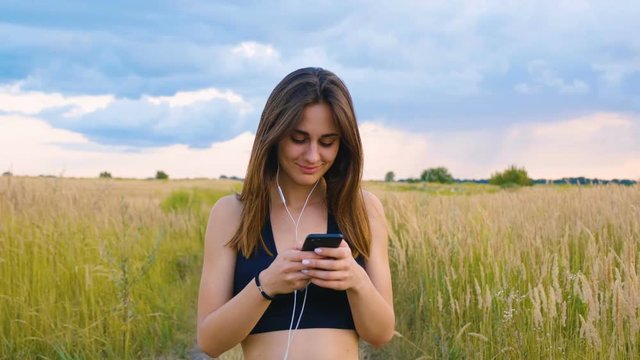 Happy young woman use smartphone and headphone walking in field at sunset, healthy lifestyle. Sportywoman runner using smartphone outdoor.
