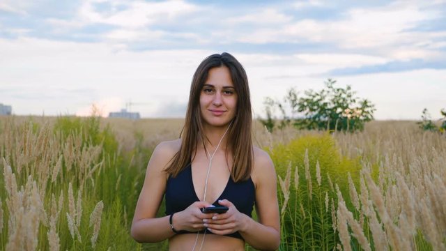 Happy young woman use smartphone and headphone walking in field at sunset, healthy lifestyle. Sportywoman runner using smartphone outdoor.