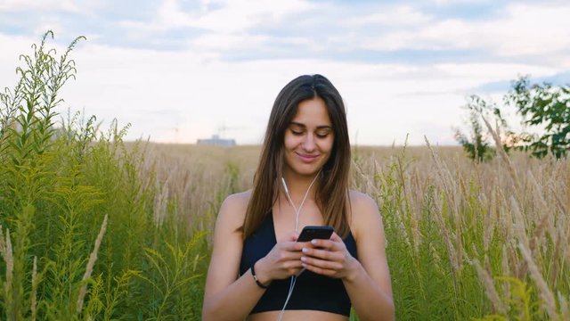 Happy young woman use smartphone and headphone walking in field at sunset, healthy lifestyle. Sportywoman runner using smartphone outdoor.