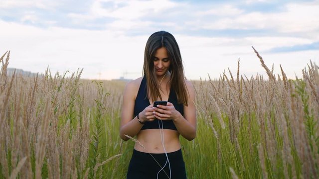 Happy young woman use smartphone and headphone walking in field at sunset, healthy lifestyle. Sportywoman runner using smartphone outdoor.