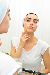Young woman, girl model,  looking at her problematic acne skin in the mirror in the bathroom,...