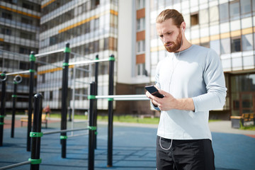 Serious concentrated young man with beard standing on workout area and using smartphone while choosing music for training