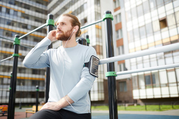 Thoughtful handsome young bearded man in sportswear standing on public sports ground and adjusting earphones while preparing for training with favorite music