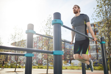 Red-bearded man in sportswear hanging on parallel bars and looking into distance while training on backyard of apartment building
