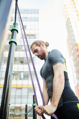 Concentrated strong young bearded man with ponytail standing at sports equipment and pulling down stretch belt