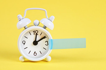 White alarm clock yellow pastel background. Blank sticky note on clock. Space copy. Minimal concept.