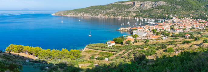 Fototapeta premium Panoramic view of a blue bay with the picturesque town of Komiza with red rooftops, white sailing boats on a summer morning viewed from above, Adriatic sea, mediterranean, Vis island, Croatia, Europe