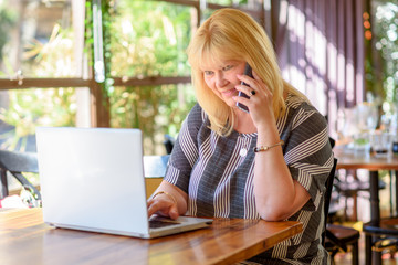 Portrait of busy sales plus size woman sitting in cafe and using her smart phone. Businesswoman working online. Attractive mature businesswoman working on laptop in her workstation.