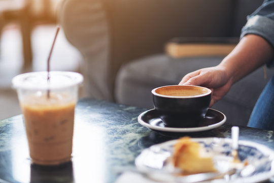 Closeup image of a hand holding a cup of hot coffee with plastic cup and dessert on table in cafe
