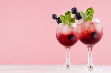 Fresh cold fruit cocktails in two elegant goblets with ice cubes, blueberry, green mint on pastel pink color background and white wood table, copy space.
