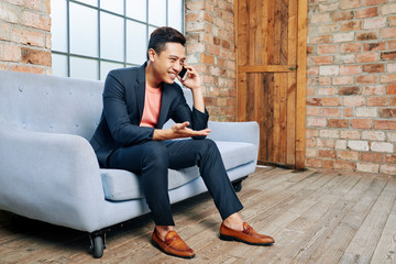 Happy young Asian entrepreneur sitting on sofa in office and explaining something coworker on the phone