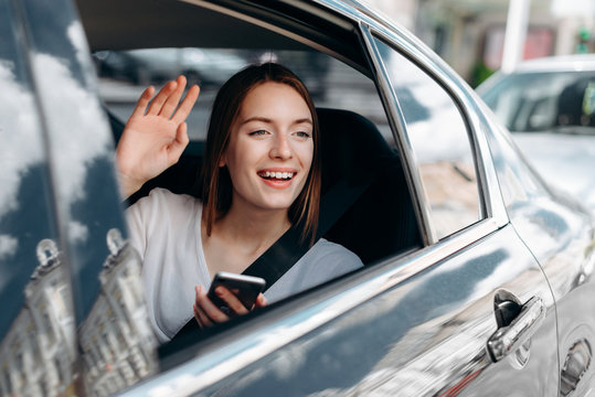 Cheerful young woman with a  smartphone sitting in the car and waving her hand look out in the window