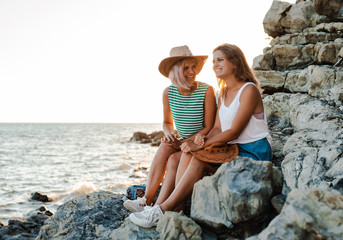 Two young cheerful women in hipsters hats on a rock on the coast of the sea. Summer landscape with girl, sea, Islands and orange sunlight.