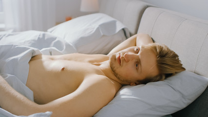 Fototapeta na wymiar Handsome Fair Boy Lies Cozily in Bed, Slowly Wakes up and Opens His Eyes. Young Caucasian Man. Early Morning Sun Shines Through the Window 