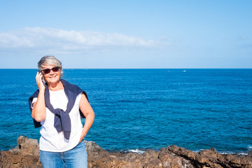 Smiling senior woman standing on the cliff over the sea. Blue sky and water. Casual clothing talking at phone. Freedom and relaxation