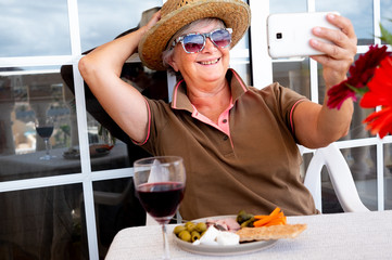 Cheerful senior woman with brown straw hat enjoying a break with red wineglass and snack. Taking a selfie with the mobile. Outdoor in the balcony looking at the mountain