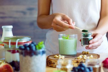 Female hands are preparing organic yogurt with spirulina for good digestion and functioning of gastrointestinal tract. Summer berries, nuts, fruits, dairy products on table. Healthy food concept.