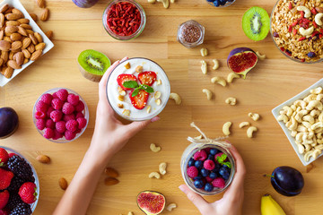 Top view on wooden table with summer breakfast, female hands are holding glass bowls with organic yoghurts. Low calorie diet meal with berries, goji, nuts, granola, chia, fruits. Healthy food concept.