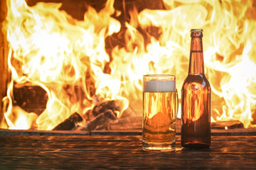 Beer in a mug and a bottle of alcohol on a wooden table on a burning fire in a fireplace background.