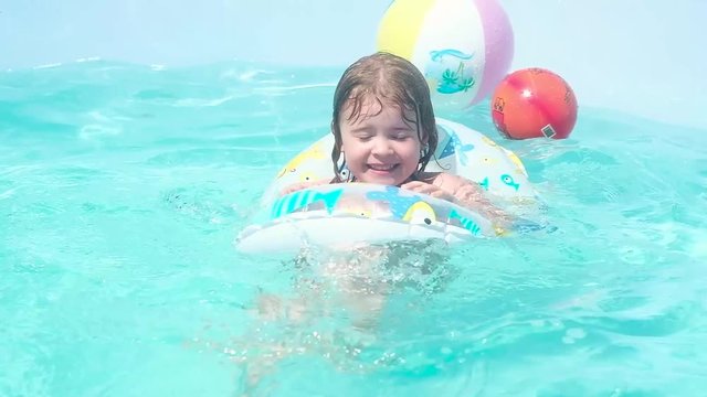 small beautiful Caucasian girl with wet dark hair swims on a children's inflatable circle in the pool. Actively pulling the legs under the water, squinting against splashing and laughing merrily.