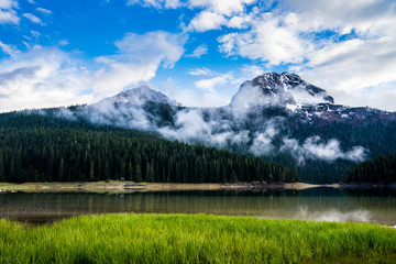 Fototapeta na wymiar Montenegro, Bobotov kuk snow covered mountains behind green forest and clouds at black lake waters in durmitor national park nature landscape near zabljak
