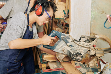 Professional young Asian female carpenter in ear muffs using circular saw when cutting pine wooden planks