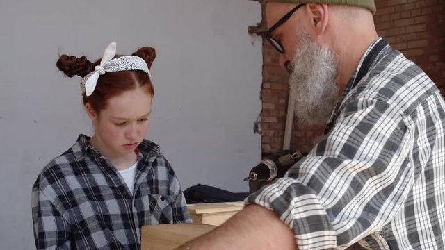 Medium shot of curious Caucasian girl watching attentively her granddad screwing screws in wooden planks with electrical hand screwdriver