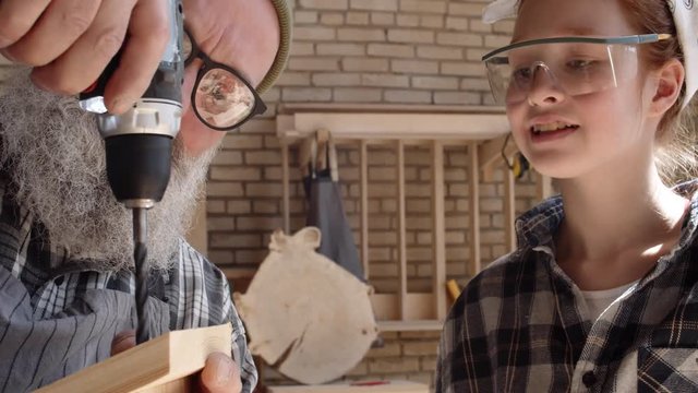 Close up of trendy joiner with beard and glasses standing at workbench with his ginger granddaughter wearing goggles and making holes in roof of wooden bird box using drill
