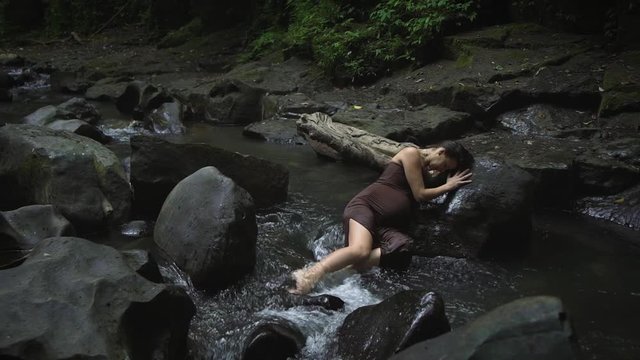 Attractive pregnant woman in a dark brown dress lies and feels labor contractions pain in the rive tropical jungle. Slow motion.