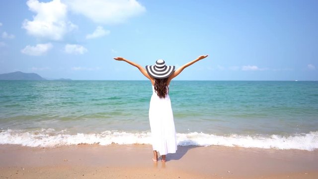 A young woman dressed in white stands on a beach, arms raised in joy.