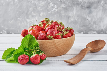 Fresh strawberries in wooden bowl with green leaves and wooden spoon on a gray background