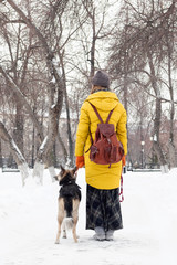 Young woman is walking in a snowy winter park with her dog. Back view.