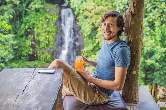 Closeup portrait image of a beautiful man drinking ice tea with feeling happy in green nature and waterfall garden background