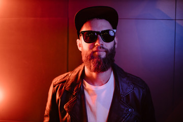Fototapeta premium Hipster handsome man on the city streets being illuminated by neon signs. He is wearing leather biker jacket or asymmetric zip jacket with black cap, jeans and sunglasses.