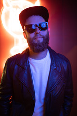 Fototapeta premium Hipster handsome man on the city streets being illuminated by neon signs. He is wearing leather biker jacket or asymmetric zip jacket with black cap, jeans and sunglasses.
