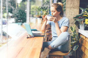 A young girl sits at the tables at the window in a cafe with a laptop, drinks matcha tea from a...