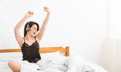 Playful woman listening to music, sitting in wireless headphones in bed