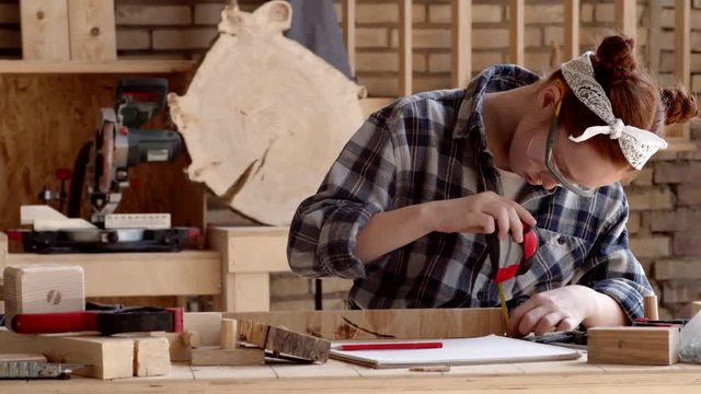 Medium shot of Caucasian red-haired girl wearing goggles standing at workbench in carpentry shop, taking measurements of wooden plank using steel tape and then writing down results with pencil