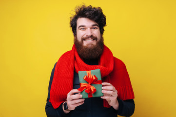 Cheerful bearded guy wearing red scarf,  smiling at camera and  holding a small gift box, standing over yellow background