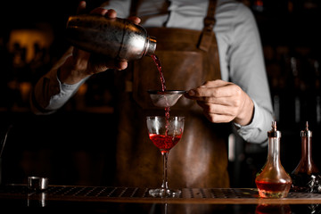 Professional male bartender pouring a red alcoholic drink from the steel shaker through the sieve