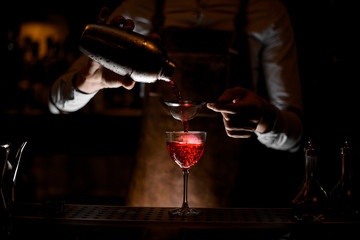 Bartender in leather apron pouring a red alcoholic drink from the steel shaker through the sieve in the dark