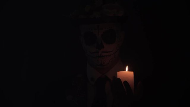 A man with his face painted as a mexican Santa Muerte skull is holding a candle