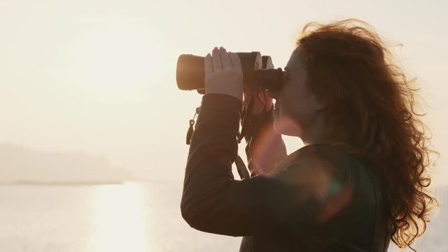 Young woman with curly hair stands on seashore at pier and looks through binoculars at sunrise and horizon of sea in the early summer morning. Light breeze puffs hair in the sun