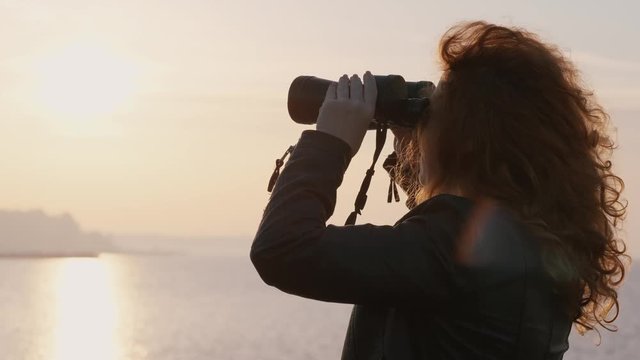 Young woman with curly hair stands on seashore at pier and looks through binoculars at sunrise and horizon of sea in the early summer morning. Light breeze puffs hair in the sun