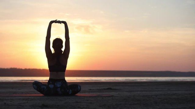 woman silhouette practicing yoga asana and improving physical and mental health sitting by sea at sunset backside view