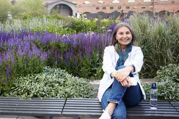 Summer image of cheerful happy middle aged woman wearing blue jeans, sneakers and white jacket sitting on bench with legs crossed, keeping hands on her knee, smiling broadly, holding mobile phone - Powered by Adobe